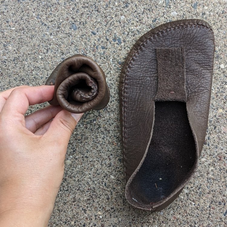 A pair of DIY Earthing Moccasins from a single piece of leather, one shoe is laying flat unworn, the other is rolled into a tight ball to show the extreme minimalist amongst barefoot shoes