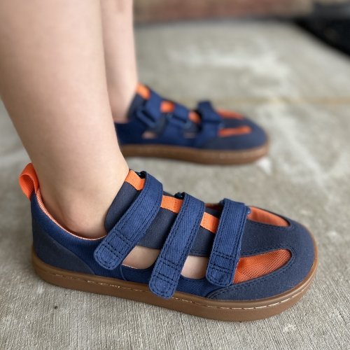 Side view of a young child wearing navy blue and orange barefoot Feelgrounds Arcade canvas sandals with adjustable straps and a wide toe box