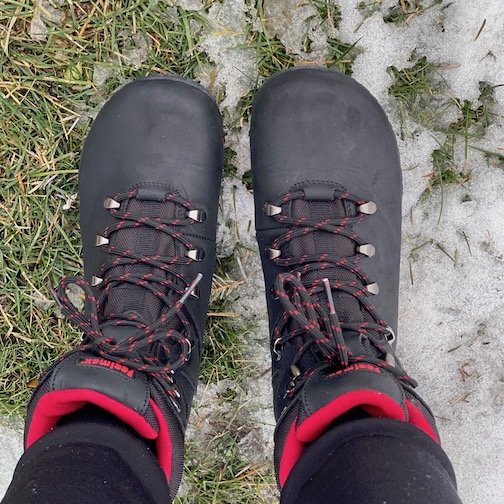 A top down view of a person wearing a pair of Feelmax Kuuva black barefoot waterproof hiking boots standing on snowy grass
