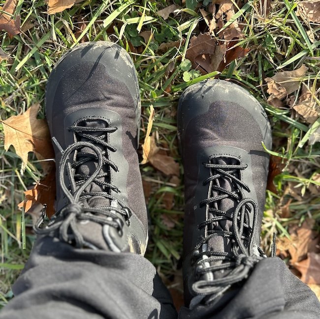 A close up top down view of a pair of feet standing in grass wearing Feet Barefoot Arken waterproof winter boots in black, showing the natural foot shape and wide toe box
