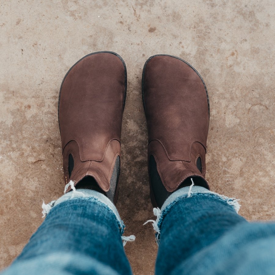 A top down view of a pair of feet standing on concrete wearing Groundies Camden in barefoot+ wide width brown leather