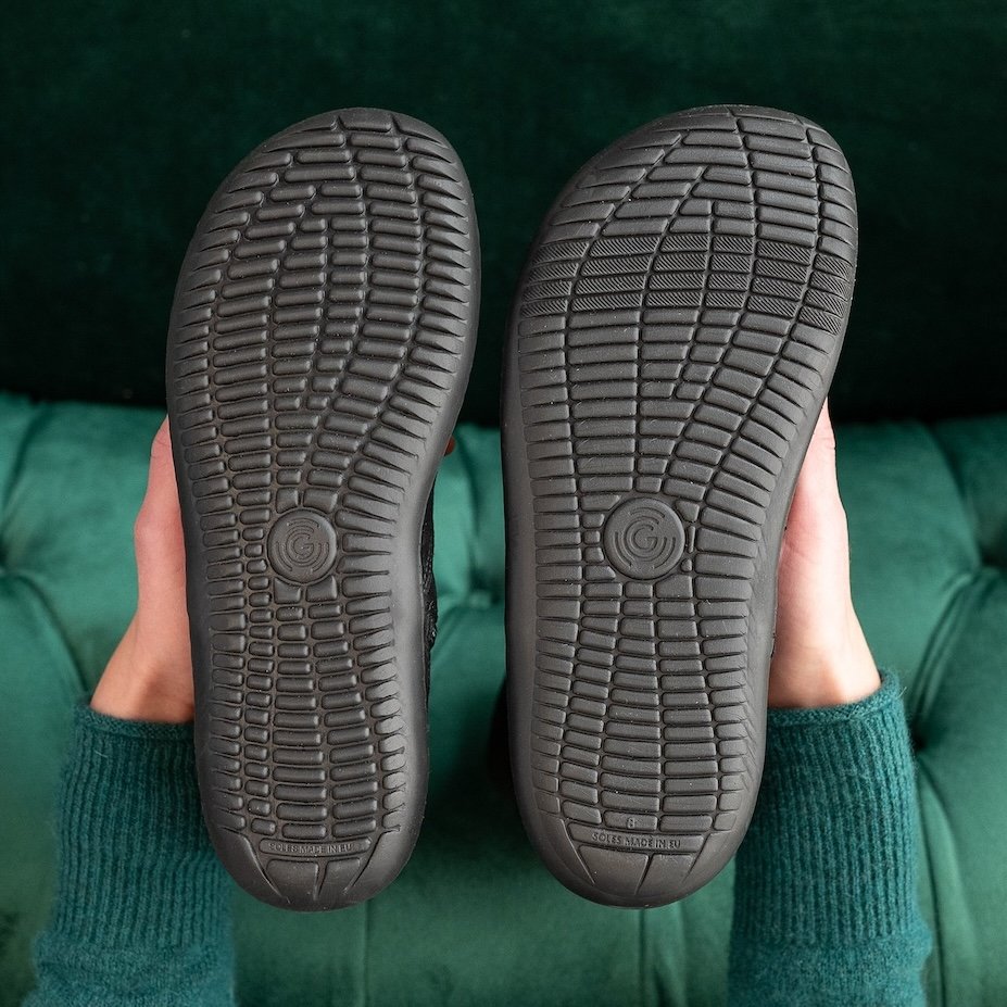 A pair of hands holding two pairs of barefoot shoes with the outsoles visible. On the left is the Groundies Barefoot width, on the right is the Groundies Barefoot Plus width, for extra wide feet