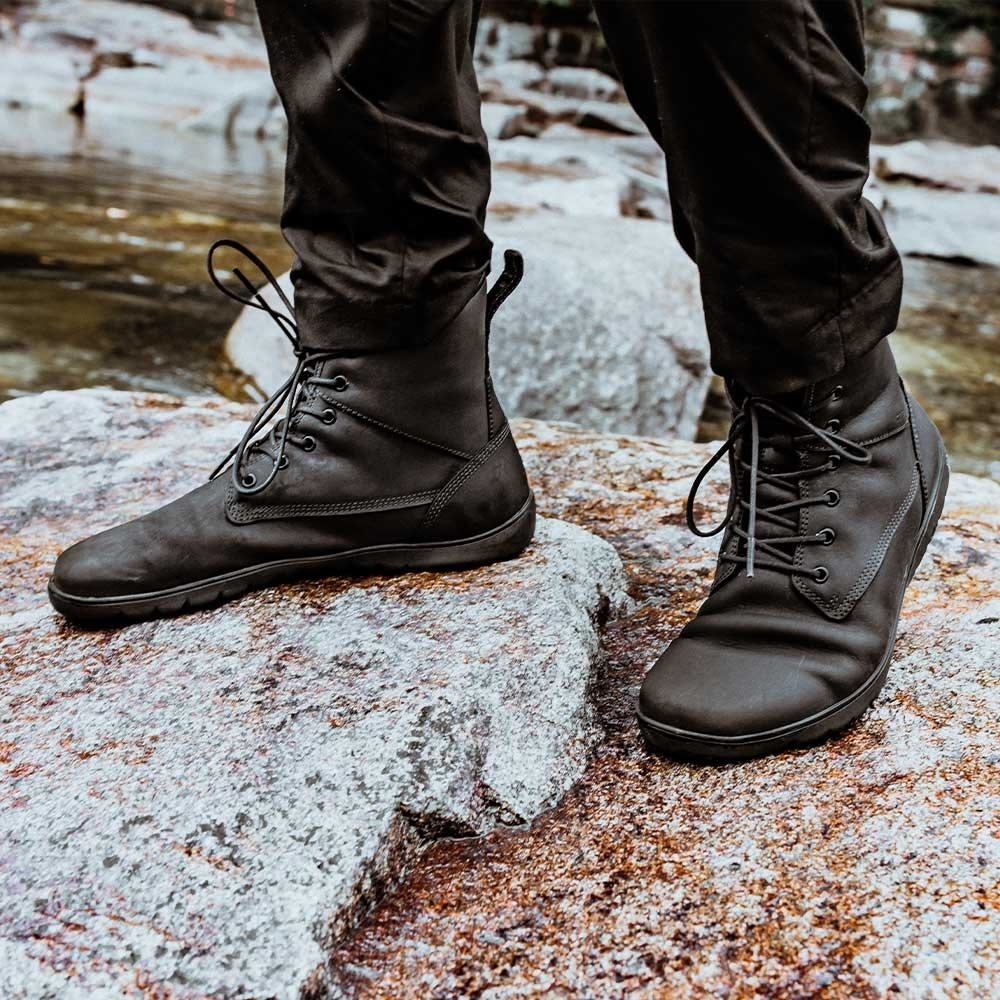A close up on a pair of man's feet wearing black pants and black Groundies Williamsburg barefoot lace up combat boots standing on rocks outdoors