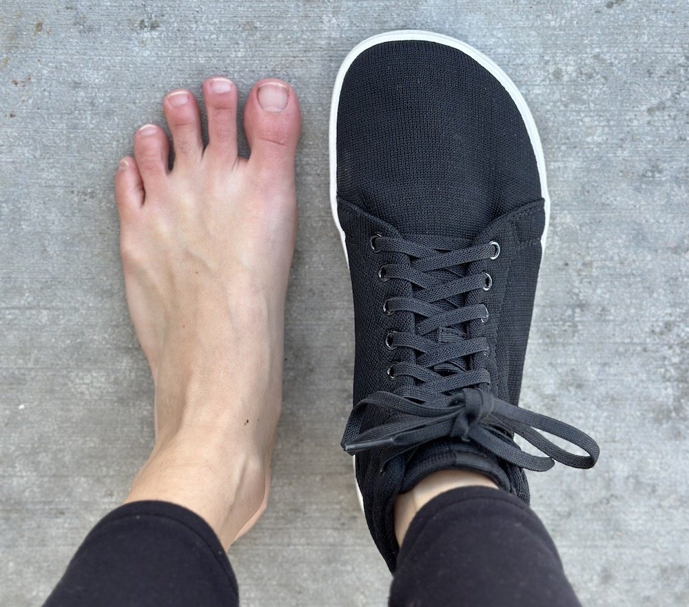 A top down view of a pair of feet standing on concrete with the left foot bare and the right foot wearing a Feelgrounds barefoot sneaker in black mesh vegan