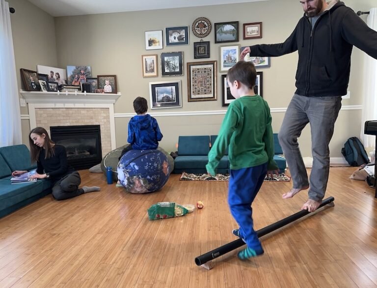 A living room with a family in it and everyone is either sitting on the floor, on cushions, or walking on balance beams