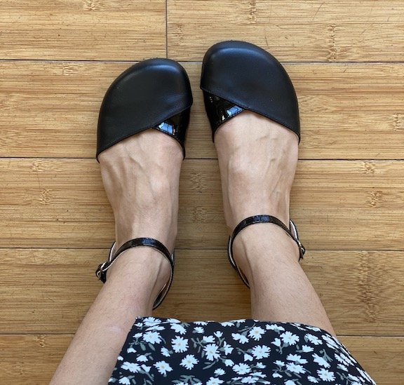 A close up of a woman's feet shown from the top wearing Shapen Poppy flats in black leather. The woman's floral skirt is visible at the bottom.