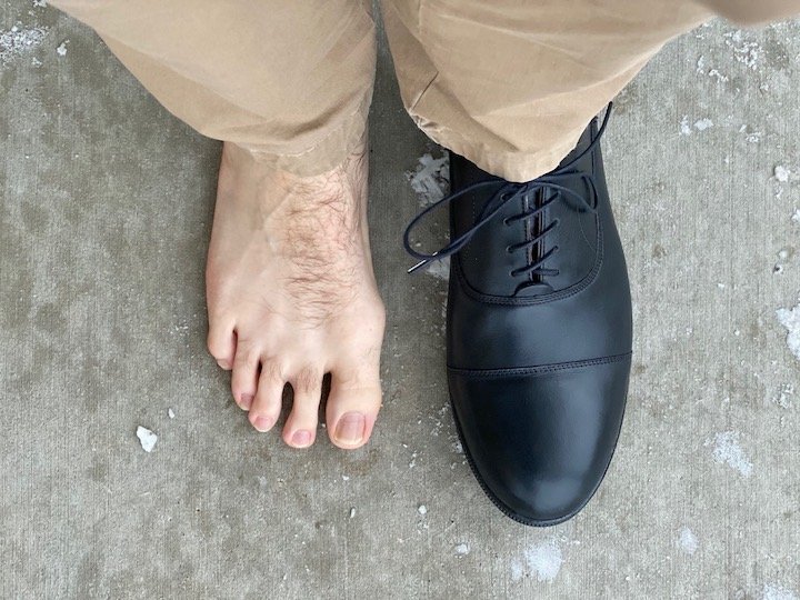 A man standing on cement with one Carets dress shoe on and one bare foot to show how the shoe tapers after the toes