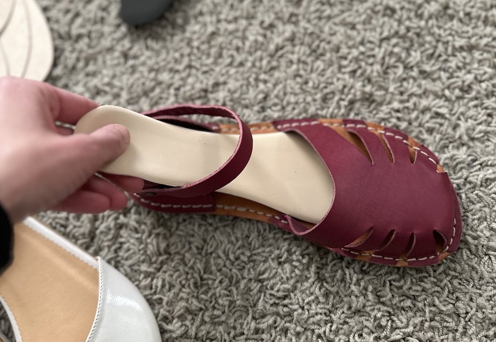 A close up of a pretty barefoot dress shoe sitting on the carpet and a hand is inserting a nude colored cushioned insole into the shoe