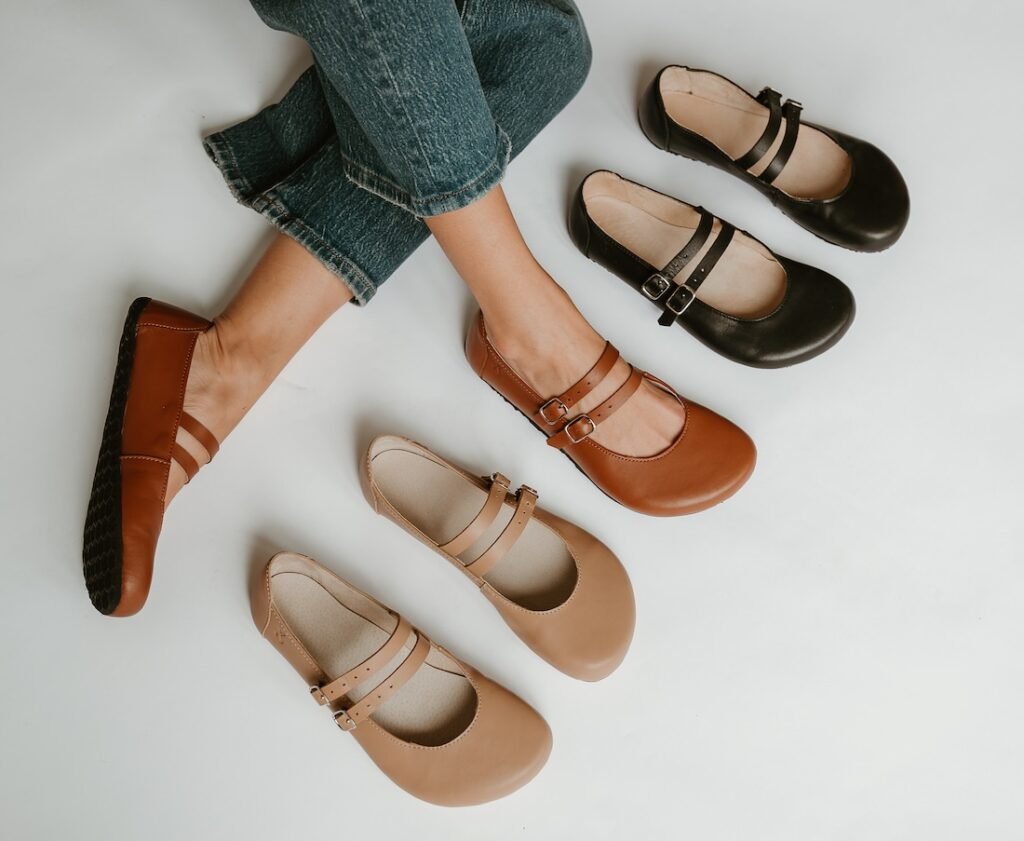 A line of Jenon Leather Villet Mary Janes with a double strap and extra wide toe box, arranged on the ground from light beige to black, with cognac in the middle being worn by a woman with her legs crossed.