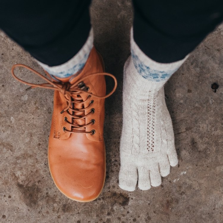 Top down view of a person wearing Knitido merino cashmere snowflake socks and a single Mukishoes cognac lace up boot on one foot with the sock peeking out the top