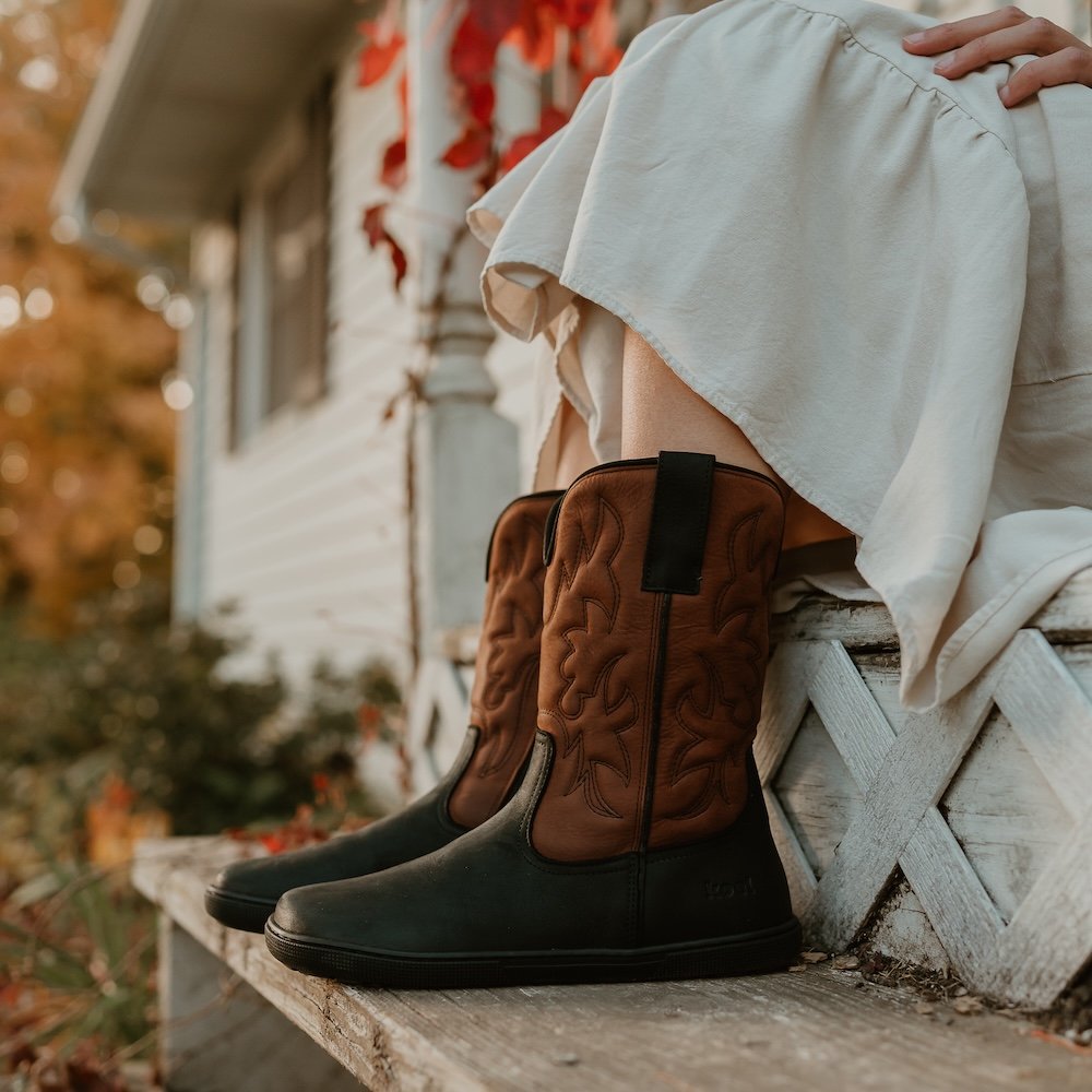 A close up side view of a person's legs and feet wearing a beige skirt and Koel Fiona black and brown leather cowboy boots with a wide toe box and flat sole