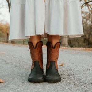 A close up of a pair of feet standing on pavement among scattered fall leaves wearing black and brown leather Koel Fiona western barefoot cowboy boots with a flat sole, wide toe box, and an embroidered pattern.