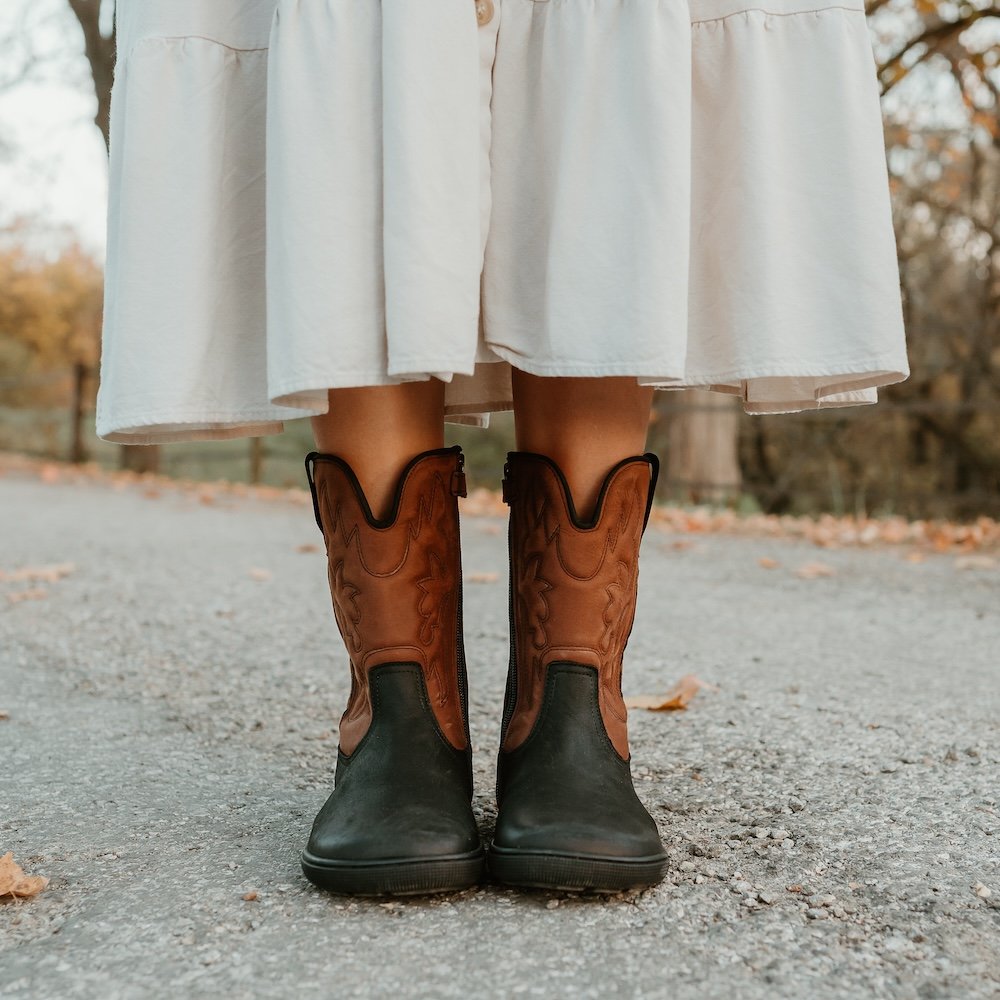 A close up of a pair of feet standing on pavement among scattered fall leaves wearing black and brown leather Koel Fiona western barefoot cowboy boots with a flat sole, wide toe box, and an embroidered pattern.