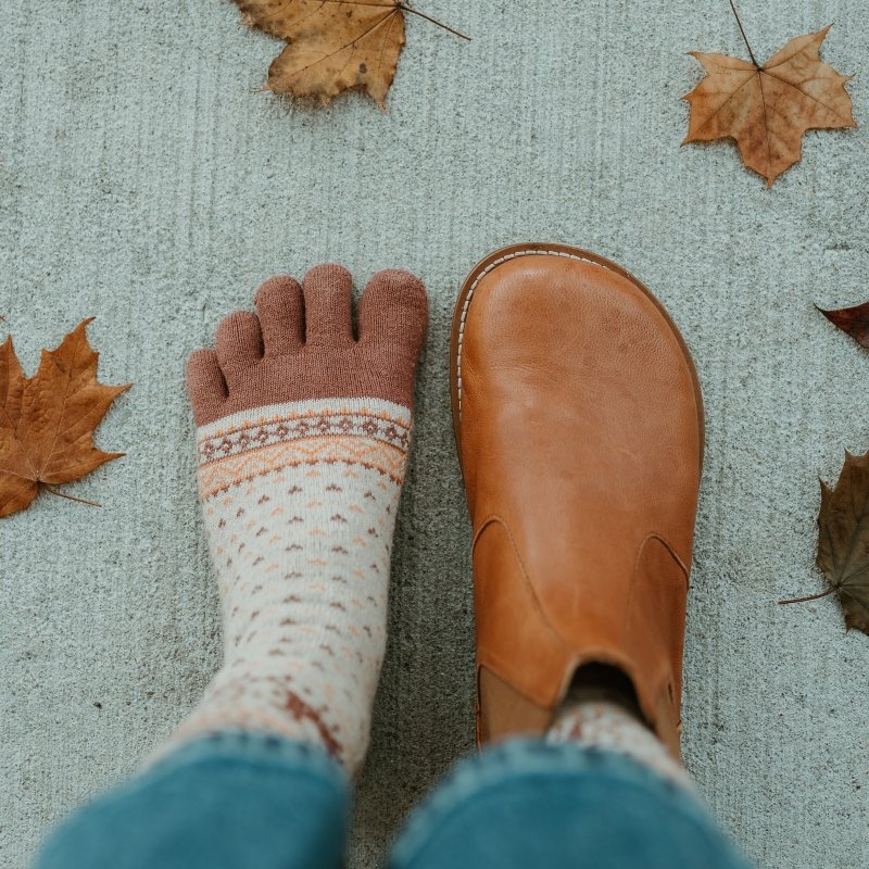 A top down view of Lang S luna caramel leather barefoot chelsea boots. The left foot is unshod and wearing a wool toe sock to show the anatomical shape match. Fall leaves are scattered against the ground.