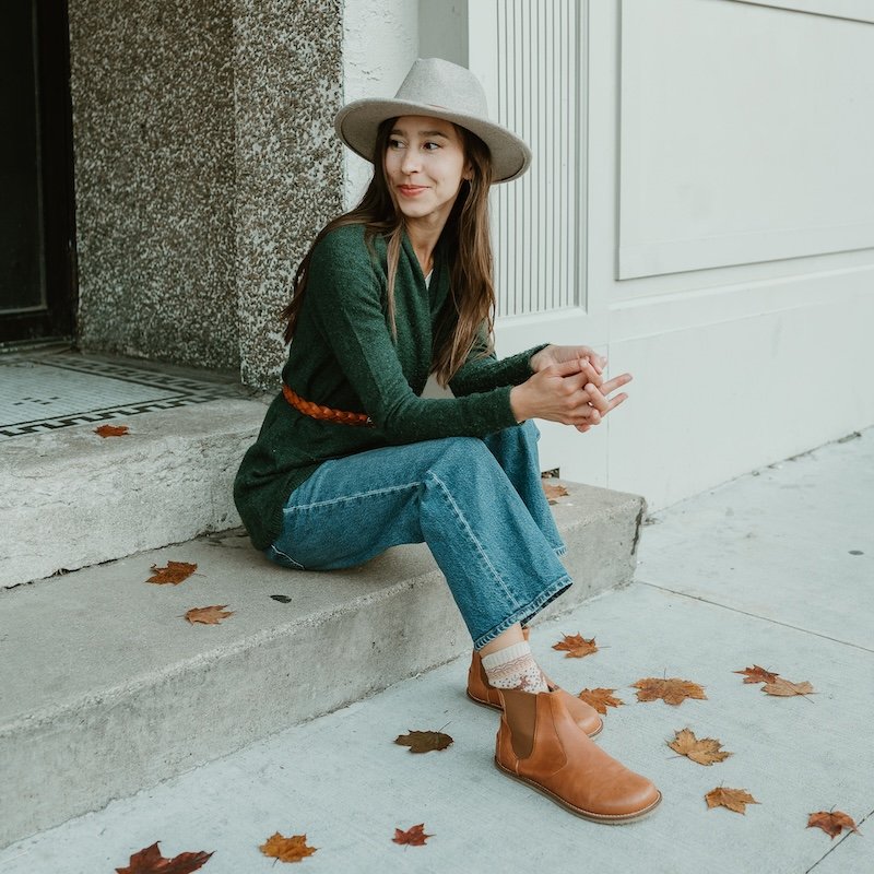 A woman sitting on a storefront steps in jeans and a dark green sweater. She also has a taupe hat and caramel leather Lang S luna barefoot chelsea boots.