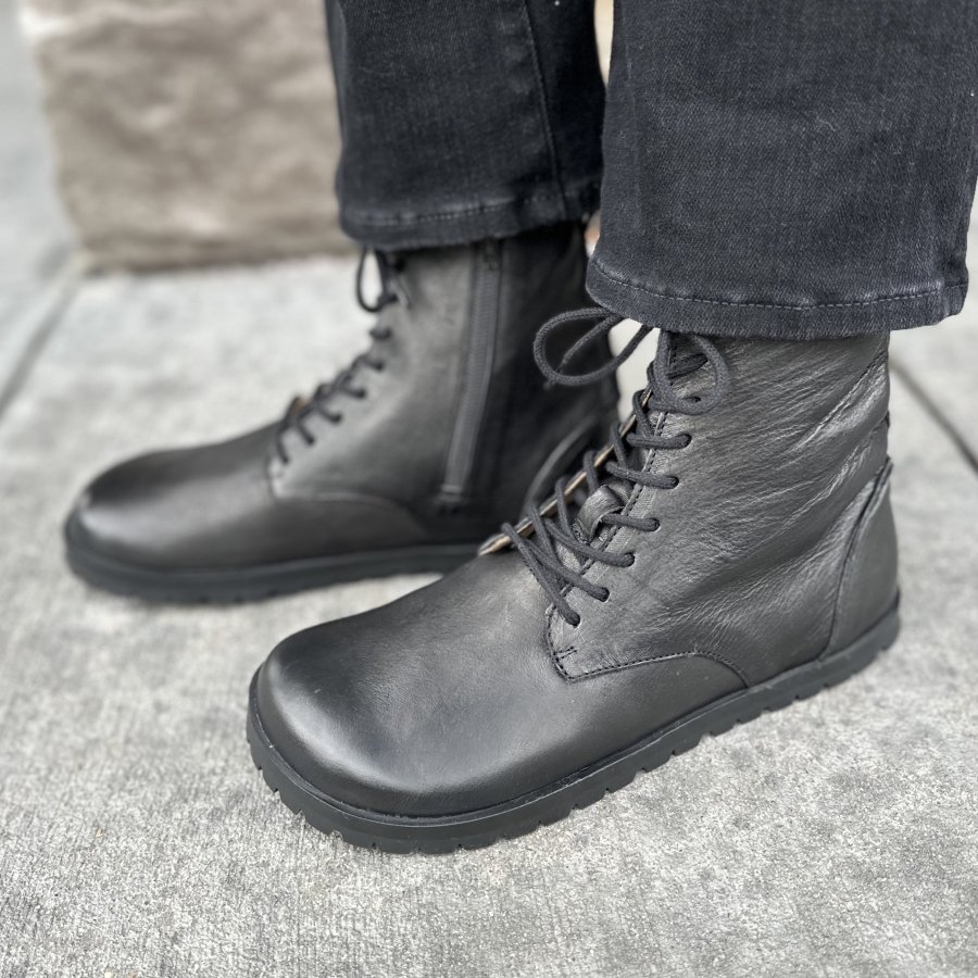 Side view of Lang S by Tikki Shoes black lace up Kombat boots with a zipper. The person is wearing them black straight jeans standing on concrete and the zero drop outsole is visible on the side