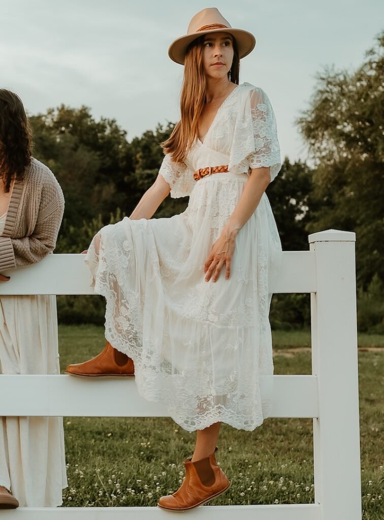 A woman sitting on a white fence outdoors wearing a white dress, cowboy hat, looking off into the distance with Lang.S Luna leather barefoot Chelsea boots on her feet