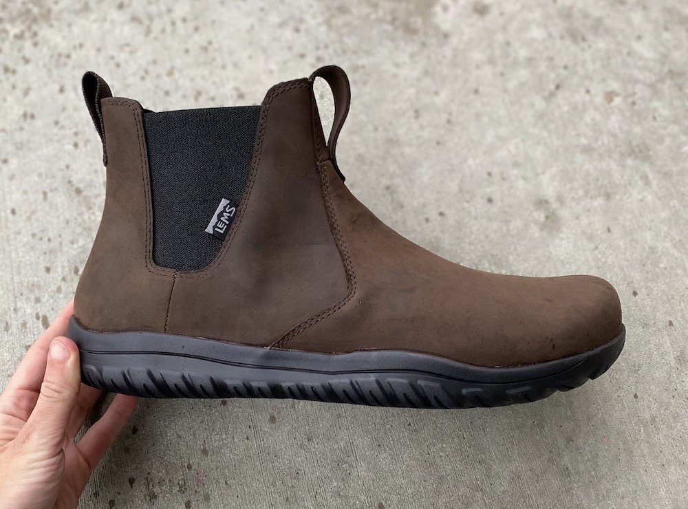 A hand holding a single Lems Chelsea waterproof brown minimalist boot similar to Blundstones