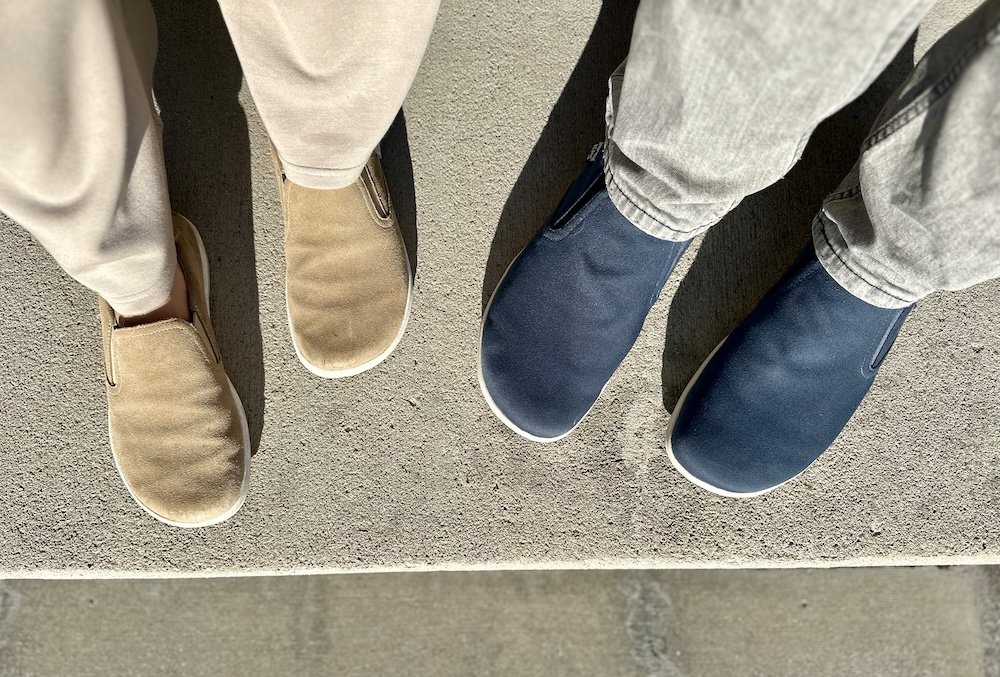 Two pairs of feet standing side by side, both wearing Lems Shoes wide toe box barefoot slip-ons, the Laguna and Malibu.