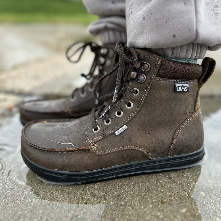 a close up side view of a pair of feet standing in a puddle in the rain wearing Dakota brown Lems barefoot waterproof Boulder boot grip with a non slip sole