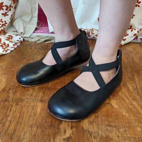 Side view of a girl's bare legs wearing black cross strap barefoot flats from LIttle Love Bug Company with a wide toe box and flexible sole