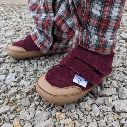 Side view of cranberry low top barefoot sneakers worn by a toddler on gravel. The reinforced leather toe edge and double velcro closure are prominent.
