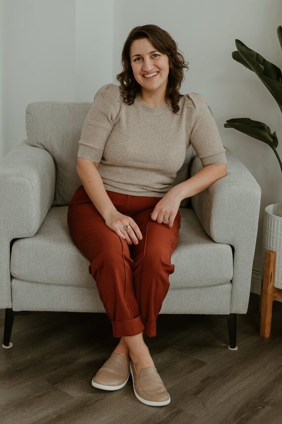 A woman with shoulder length wavy brown hair sitting in a chair with her feet crossed. She has neutral Be Lenka Buena barefoot loafers on her feet, rust colored dress pants, and a tan beige sweater