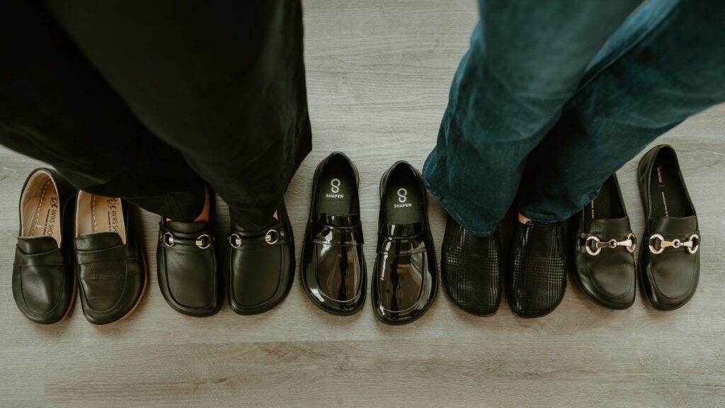 A top down view of two women standing in different pairs of black loafers amount a line of all black barefoot loafers