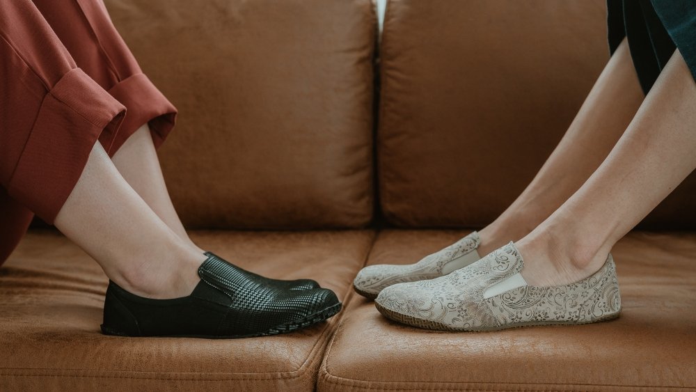 2 women's feet in Peerko Trim loafers resting on a couch facing eachother. They are each showing Trim barefoot laofers in tan paisley and black plaid