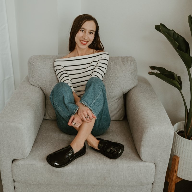A woman in a white shirt with black stripes sitting on a chair. her ankles are crossed and she has Sapen Sheen patent leather barefoot loafers on her feet. She is smiling at the camera with her hands resting on her legs.