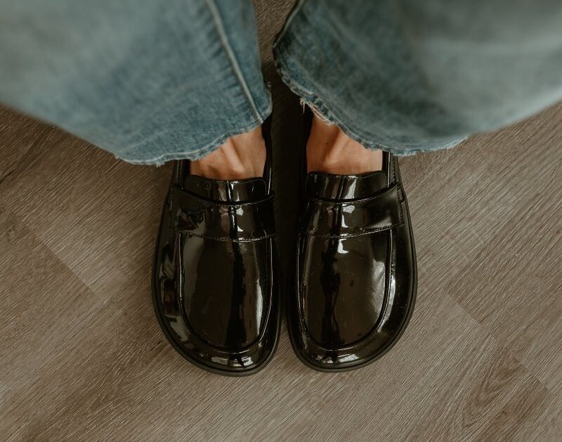 Top down view of Shapen Barefoot Sheen wide to box patent leather loafers for comfortable all day wear. There are a pair of mid tone denim showing above the shoes.