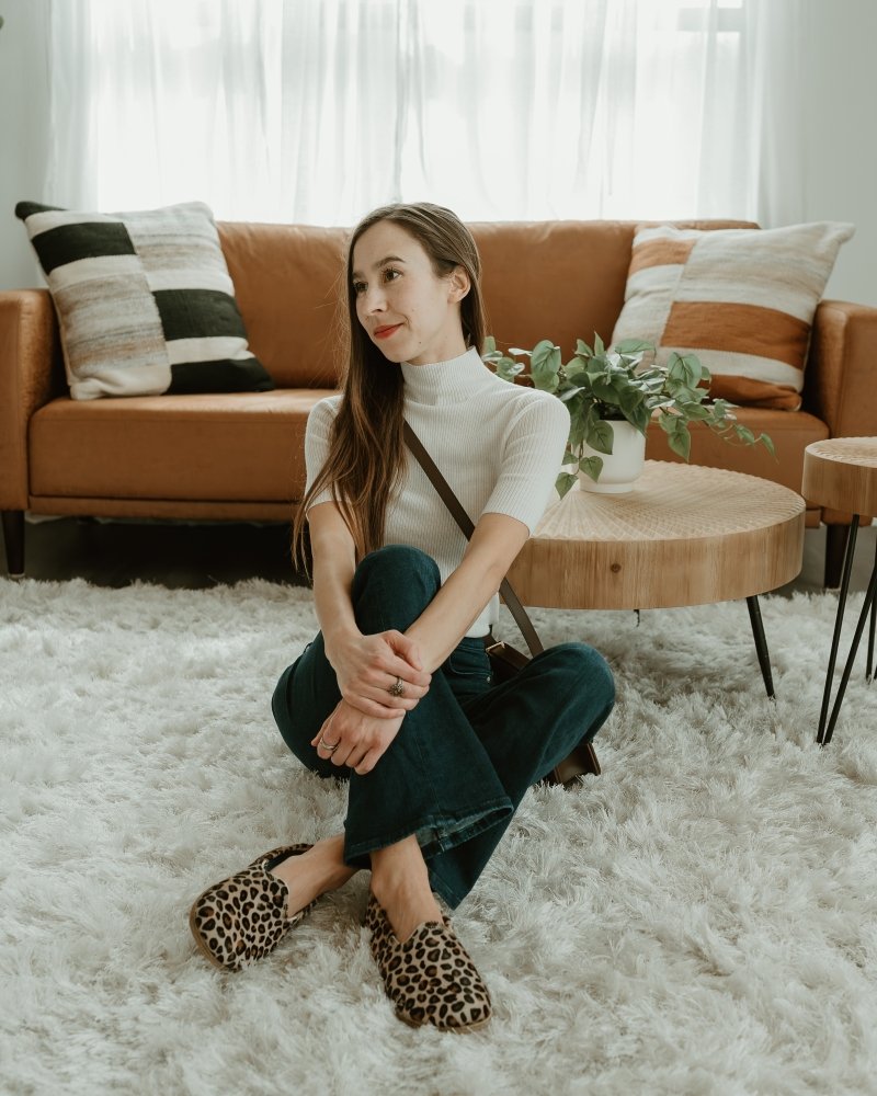 A woman with long brown hair wearing a white mock neck sweater and flare leg jeans sitting cross legged on the floor in Anya's Shop Yasemin Loafers in leopard