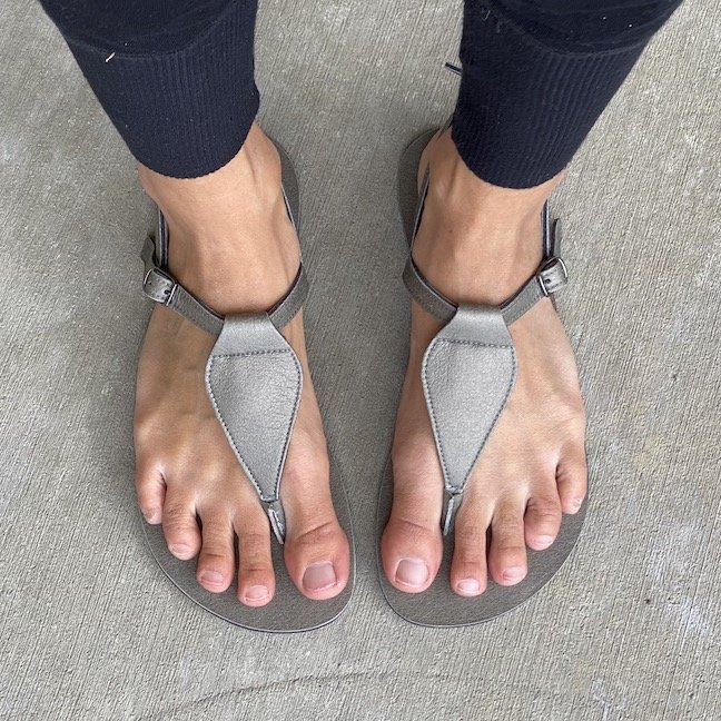 A top down view of a pair of feet standing on concrete wearing silver metallic Lonnies - stylish barefoot sandals for women
