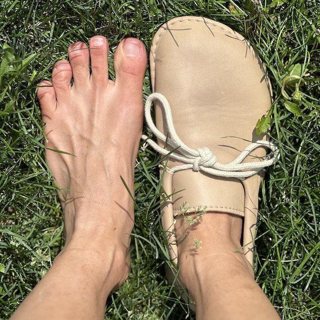 Luks Cordelia barefoot slip ons moccasins handmade on a pair of feet standing on grass, with the left foot bare to show that the shoe is shaped like a natural foot