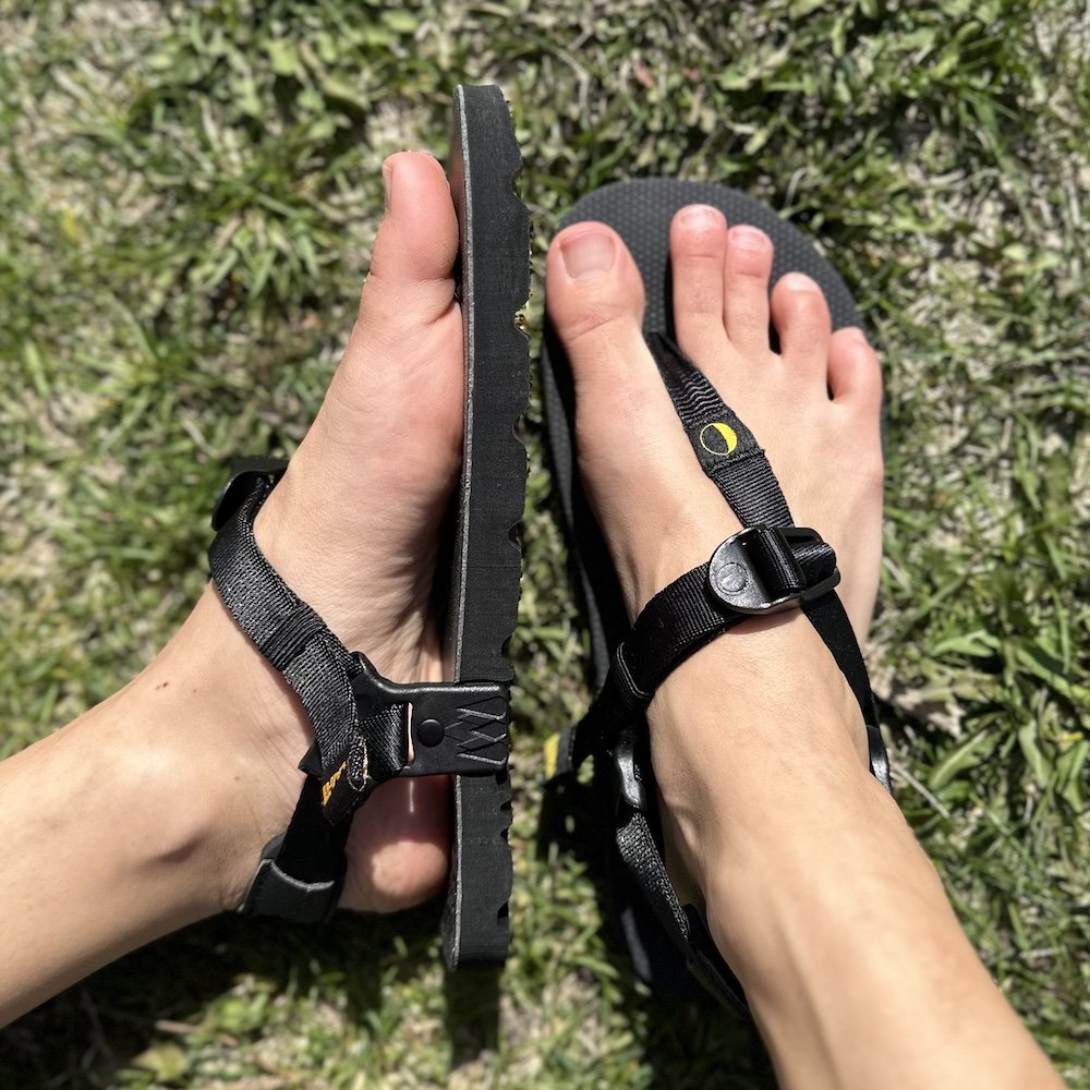 A close up of a pair of feet resting on grass wearing Luna Barefoot sandals Mono in black