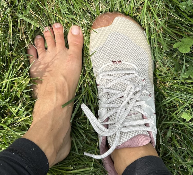 A top down view of a pair of feet standing on grass. One foot is bare and the other is wearing a Merrell Vapor Trail Glove barefoot hiking shoe