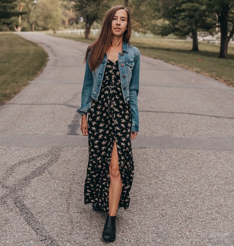 A woman walking down a curved road with trees in the background showing how she wears Mukishoes Chelsea black leather barefoot boots for fall. Her long black floral dress has a slit mid-thigh and she has paired them with a denim jacket.