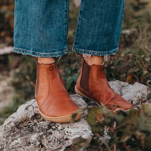 A pair of legs wearing Mukishoes Chelsea in Caramel leather, sustainable barefoot boots outside on a white rock with green foliage in the back and foreground.