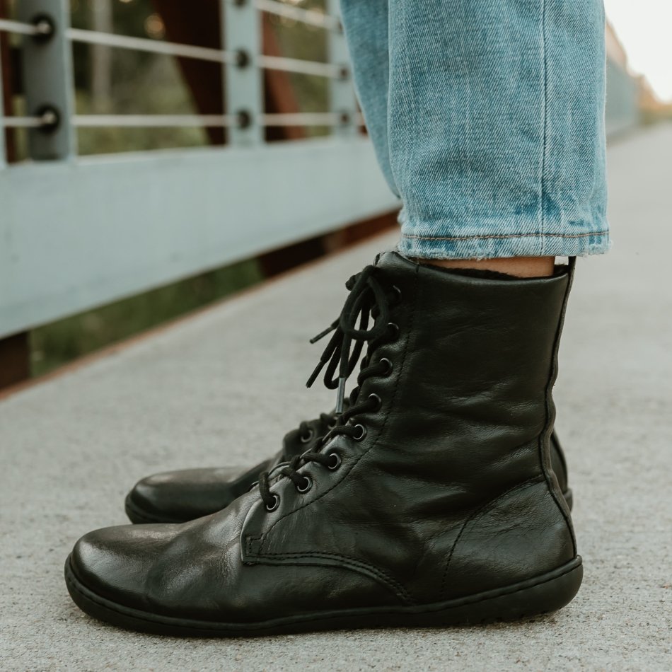 Close up side view of black barefoot wide toe box combat boot style leather boots with wool lining. The Mukishoes Igneous boots are shown worn with straight leg jeans outside on a bridge