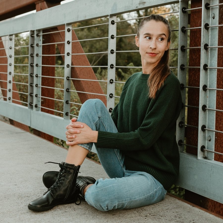 A woman sitting on a bridge with her back against the side looking off camera with a slight smile. She is wearing a dark green sweater, light colored slim jeans, and Black leather combat style lace up barefoot Igneous boots from Mukishoes.