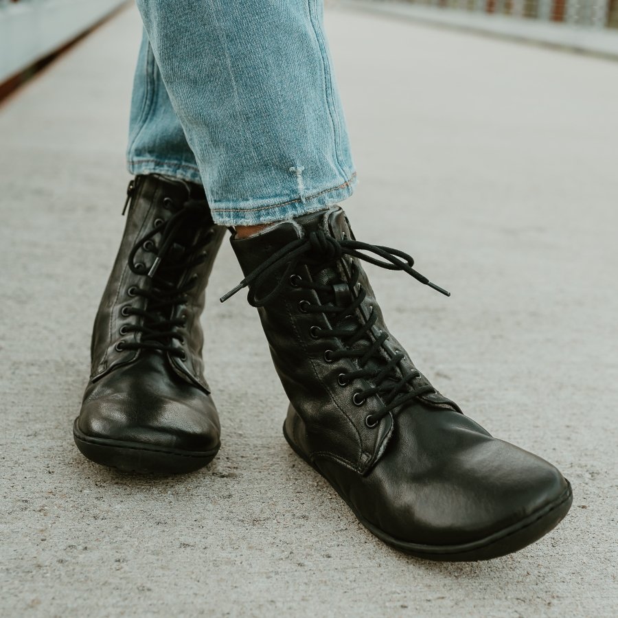 Close up view of a pair of feet crossed on the sidewalk. The person is wearing slim light colored jeans with black lace up wool lined leather boots from Mukishoes. The sustainable barefoot Igneous boots have laces and a zipper.