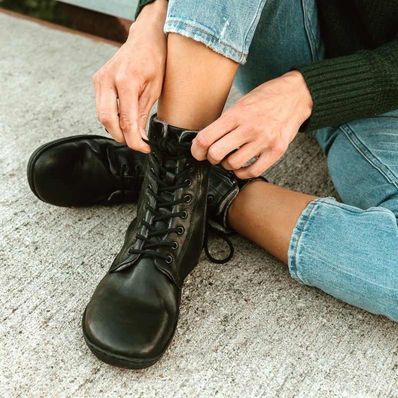 Close up shot of a person sitting on the ground with ankles crossed wearing a chunky green sweater, light wash jeans, and black leather wool lined lace up barefoot boots from Mukishoes. The Igneous model has a zipper visible int he background and laces shown in the front for an adjustable fit. The wide toe box is prominently shown in the front of the image