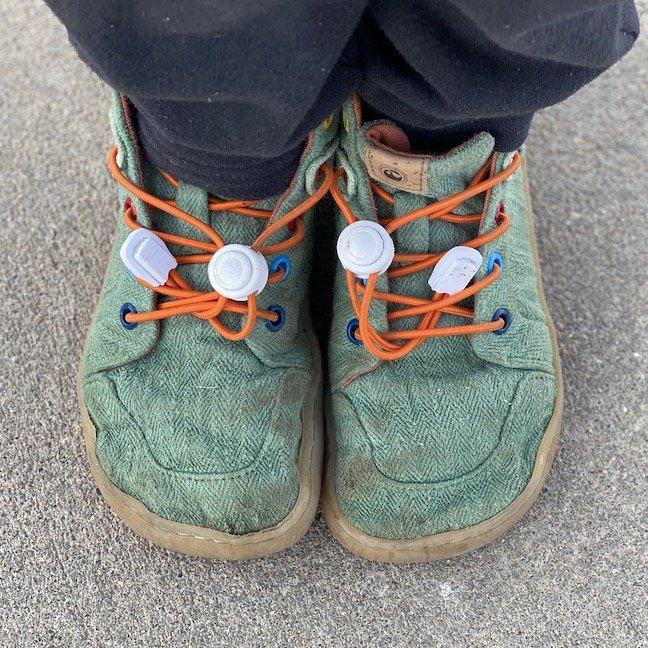 A top down view of a child's feet wearing Mukishoes Mini Moss green barefoot shoes for kids with orange laces and rainbow eyelets to show the wide anatomical toe box shape