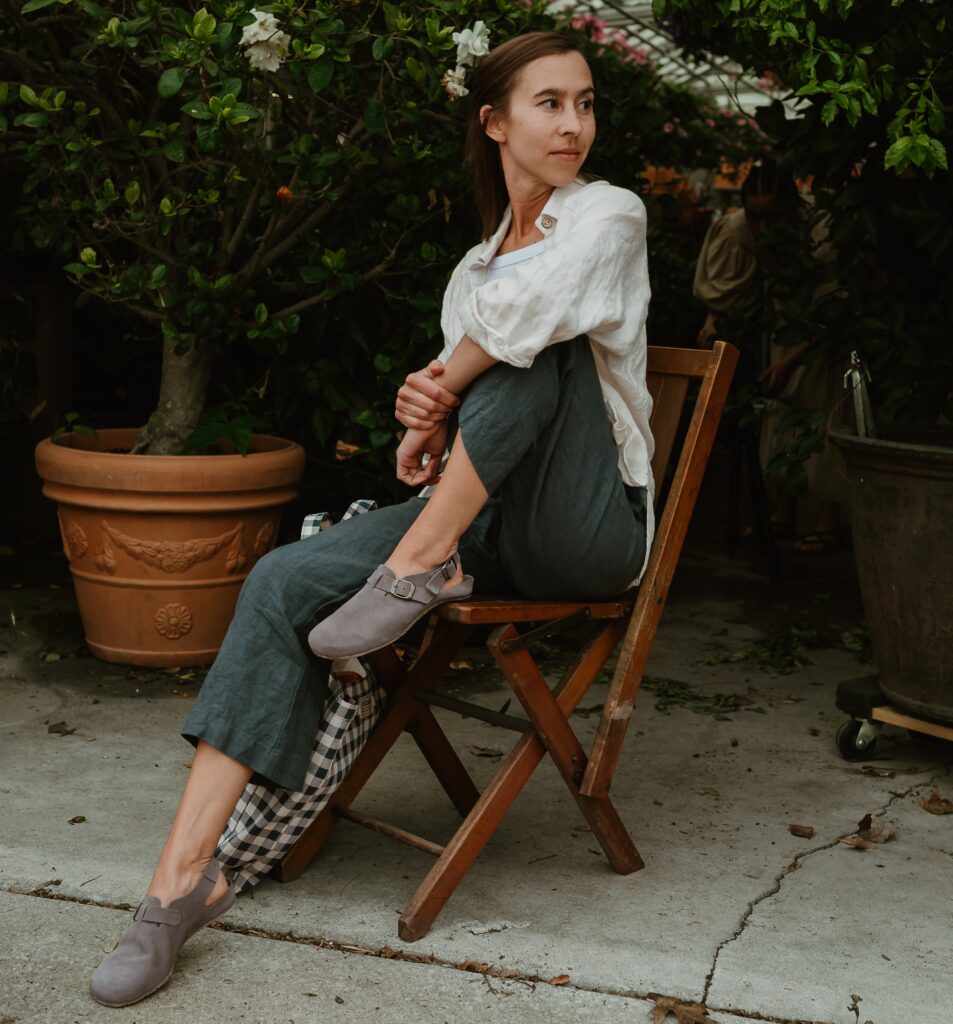Full body view of a woman sitting on a wooden folding chair wearing dark gren linen pants and a breezy linen button up. She is holding one knee close to her body and looking off into the distance. On her feet are comfortable wide toe box flexible Soca clogs from Mukishoes