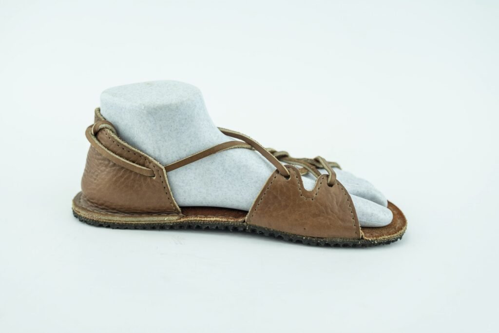 A tan leather barefoot sandal with a heel cup and thick straps over the forefoot. Leather on heels and forefoot is attached by thin leather straps. Shown on a white background.