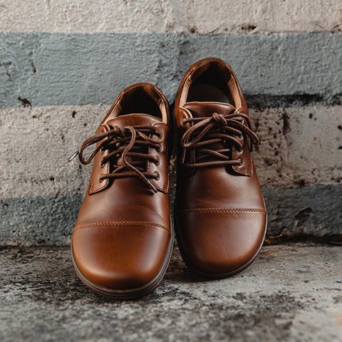 The Lems Shoes Nine2Five men's wide toe box barefoot dress shoes in brown leaning against a cement wall