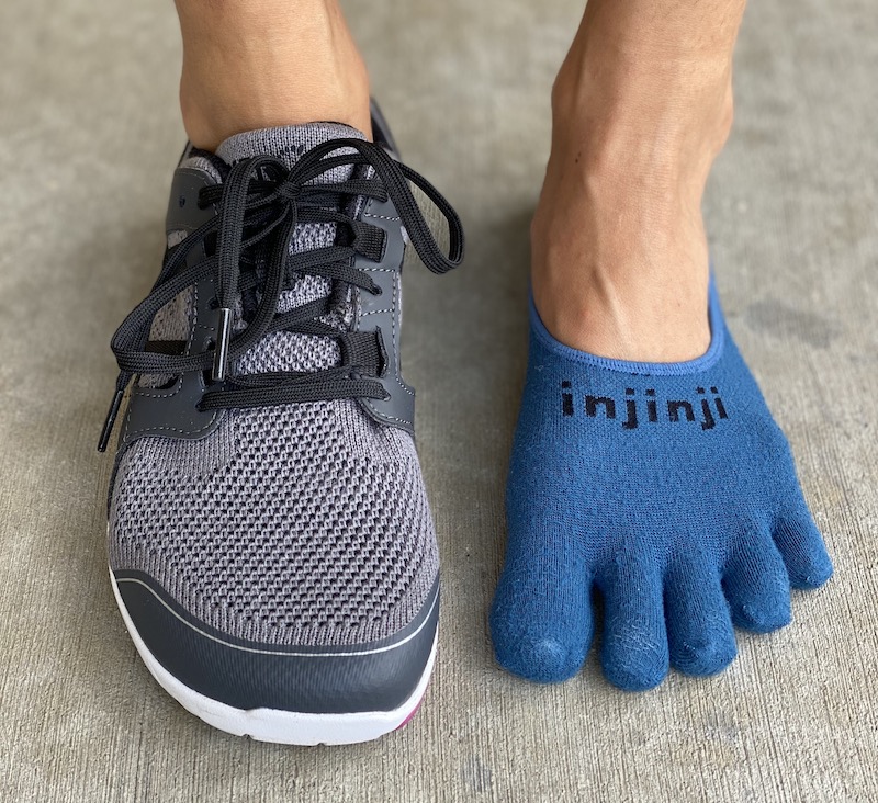 A close up of a pair of feet standing on concrete. The right foot is wearing a Xero Shoes Zelen barefoot sneaker, the left foot is wearing an Injinji hidden toe sock