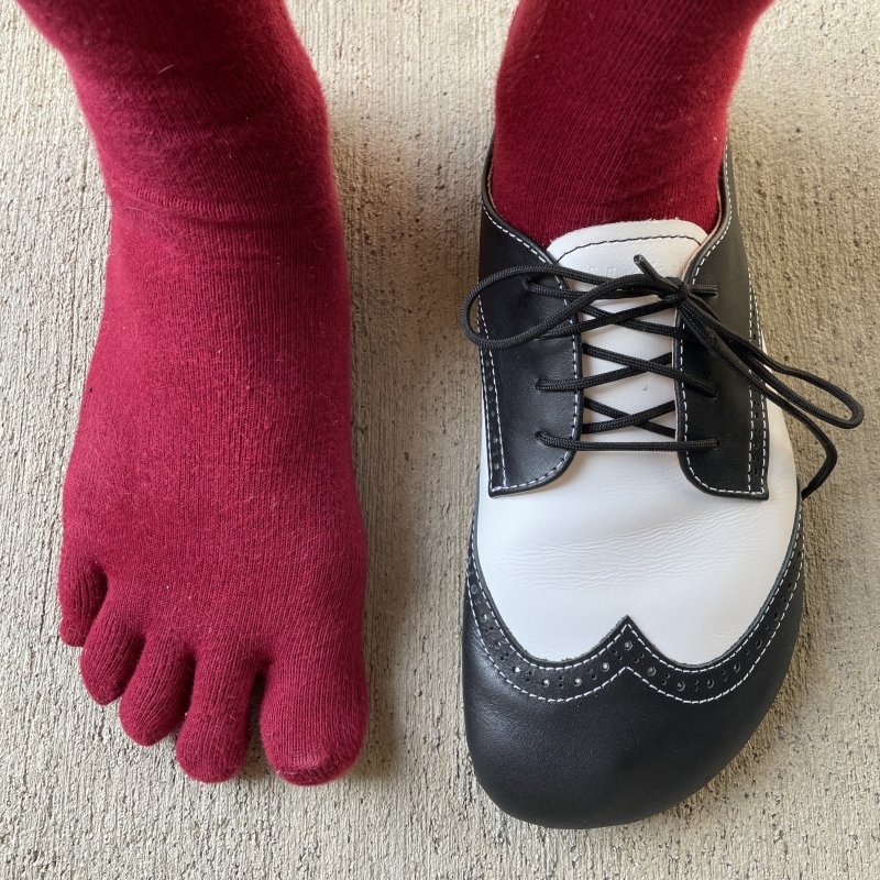 Top down view of a foot with red toe socks on wearing a black and white leather saddle style barefoot shoe from NoNs