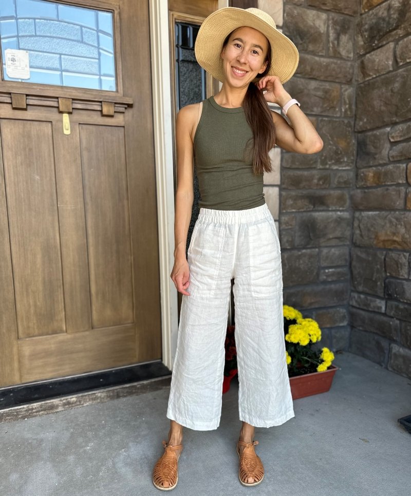 A woman in a summery outfit featuring Not Perfect Linen Puglia Pants, an olive tank top, straw hat, and Origo Huarache Sandals by Anya