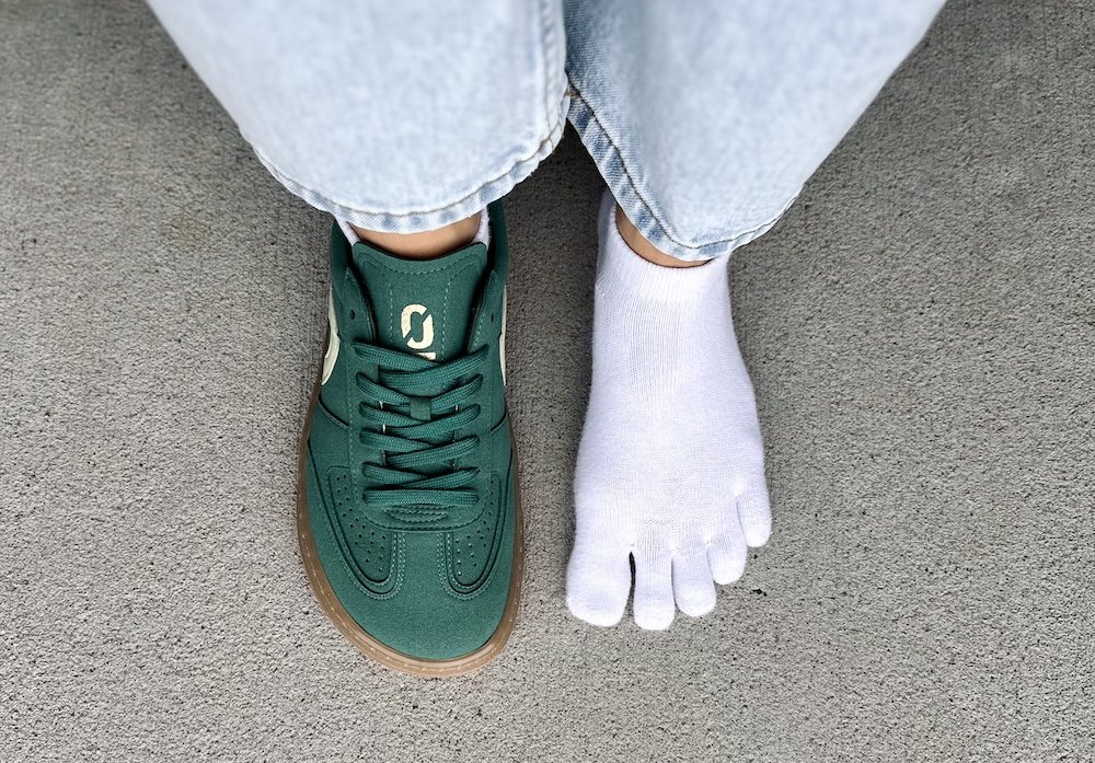 A top down view of a pair of feet. One foot is wearing toe socks and the other is wearing an Ohne Project vegan barefoot sneaker that is dark green